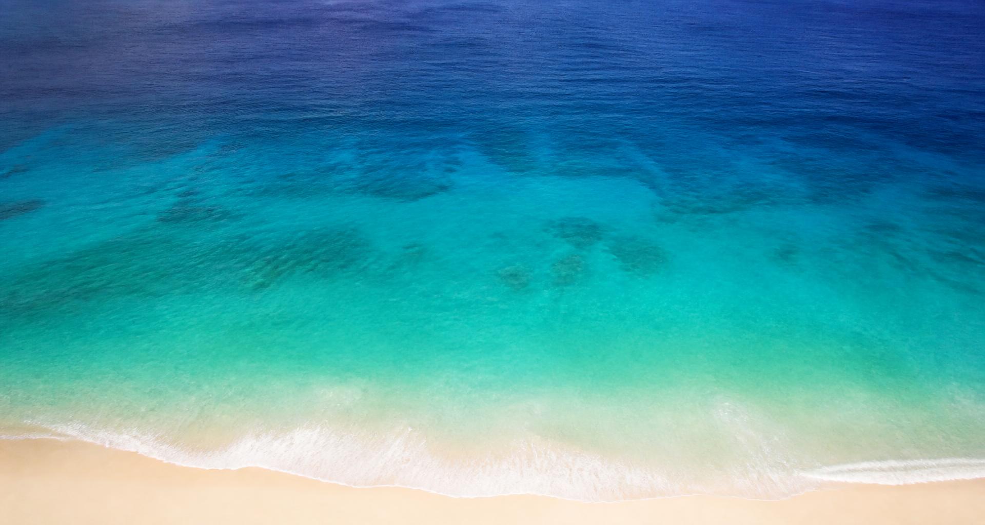Aerial view of a beach where turquoise shallows deepen into dark blue ocean